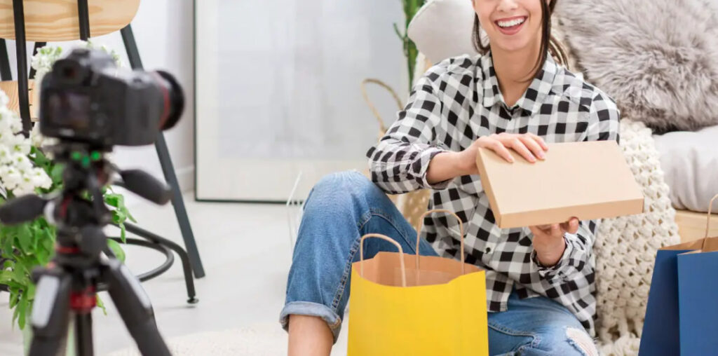 How to do product placement: a woman sitting on the floor, holding a camera and shopping bags, smiling enthusiastically.