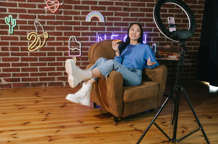 Une femme assise sur une chaise, tenant un appareil photo, avec un panneau néon illuminé en arrière-plan.